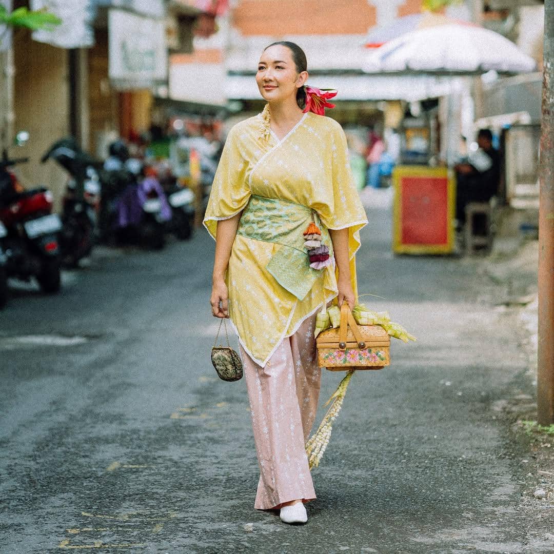 Perempuan mengenakan kebaya kuning dan kain batik berjalan di jalan kecil sambil membawa tas anyaman dan sesajen di tangannya.