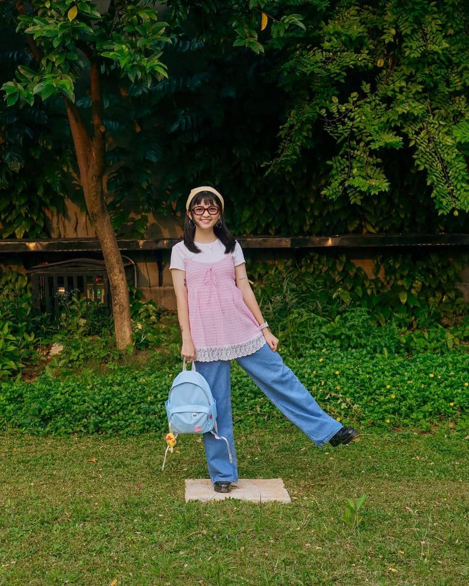 Seorang perempuan bergaya kasual mengenakan atasan merah muda, celana jeans longgar, dan membawa ransel biru muda di taman hijau.