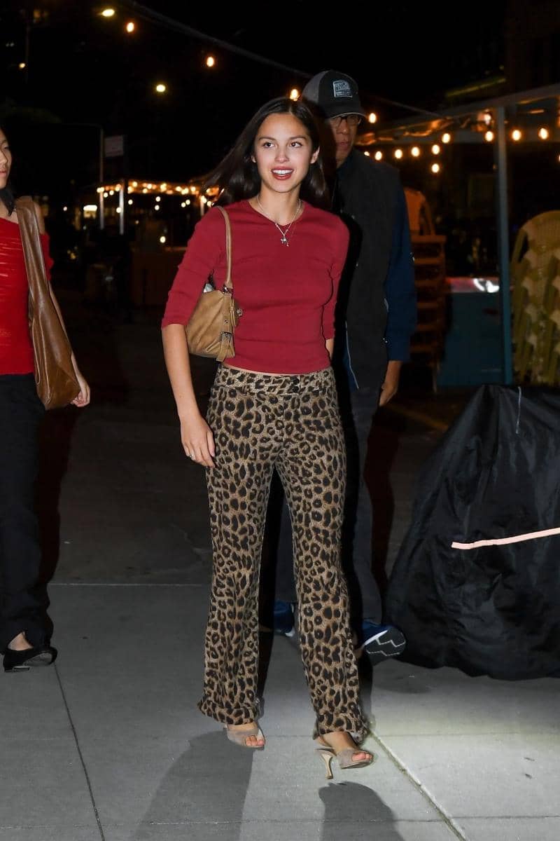 Seorang wanita mengenakan atasan merah dan celana bermotif leopard berjalan di trotoar pada malam hari dengan lampu hias di latar belakang.
