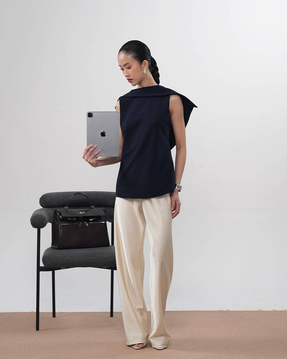 Seorang wanita berpose memegang tablet di tangan, mengenakan atasan tanpa lengan berwarna hitam dan celana panjang krem di ruangan minimalis.