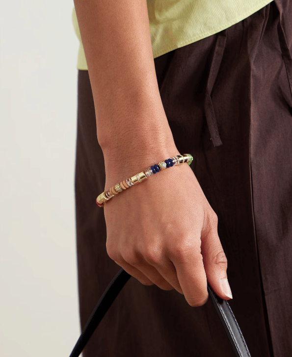 Gelang manik-manik berwarna emas, biru, hijau, dan merah dikenakan di pergelangan tangan seseorang yang memegang tali tas hitam.