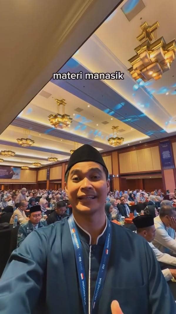 Seorang pria mengenakan baju koko biru dan peci hitam berfoto di aula besar bersama peserta lain yang mengikuti kegiatan manasik haji.