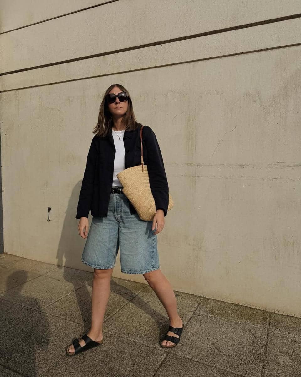 Seorang wanita mengenakan kemeja hitam, kaus putih, celana pendek denim, dan sandal hitam sambil membawa tas anyaman besar di bahunya.