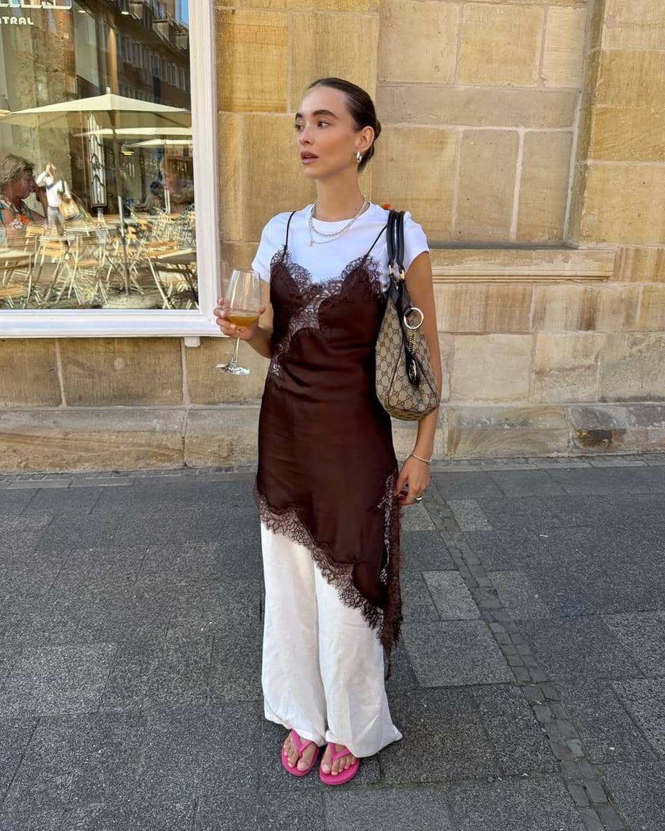 Seorang wanita berdiri di jalan mengenakan gaun cokelat berenda di atas kaus putih, celana panjang putih, dan sandal jepit merah muda sambil memegang gelas minuman.