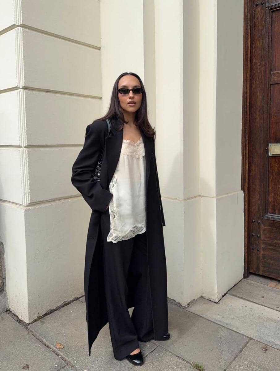 Seorang wanita bergaya chic mengenakan mantel panjang hitam, blus satin putih, dan celana lebar hitam di depan bangunan klasik.