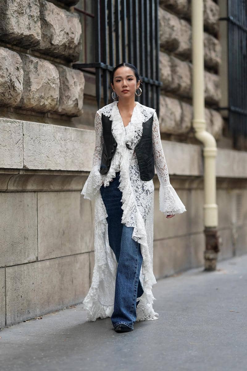 Seorang wanita berjalan di trotoar mengenakan blus renda putih panjang dengan rompi hitam dan celana jeans biru bergaya kasual elegan.