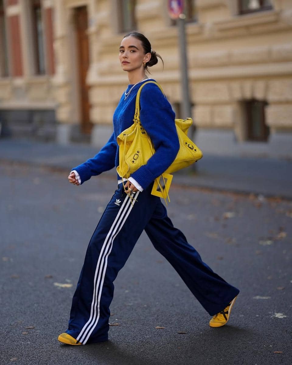 Seorang wanita berjalan di jalan kota mengenakan sweater biru, celana olahraga biru bergaris putih, dan tas serta sepatu kuning.