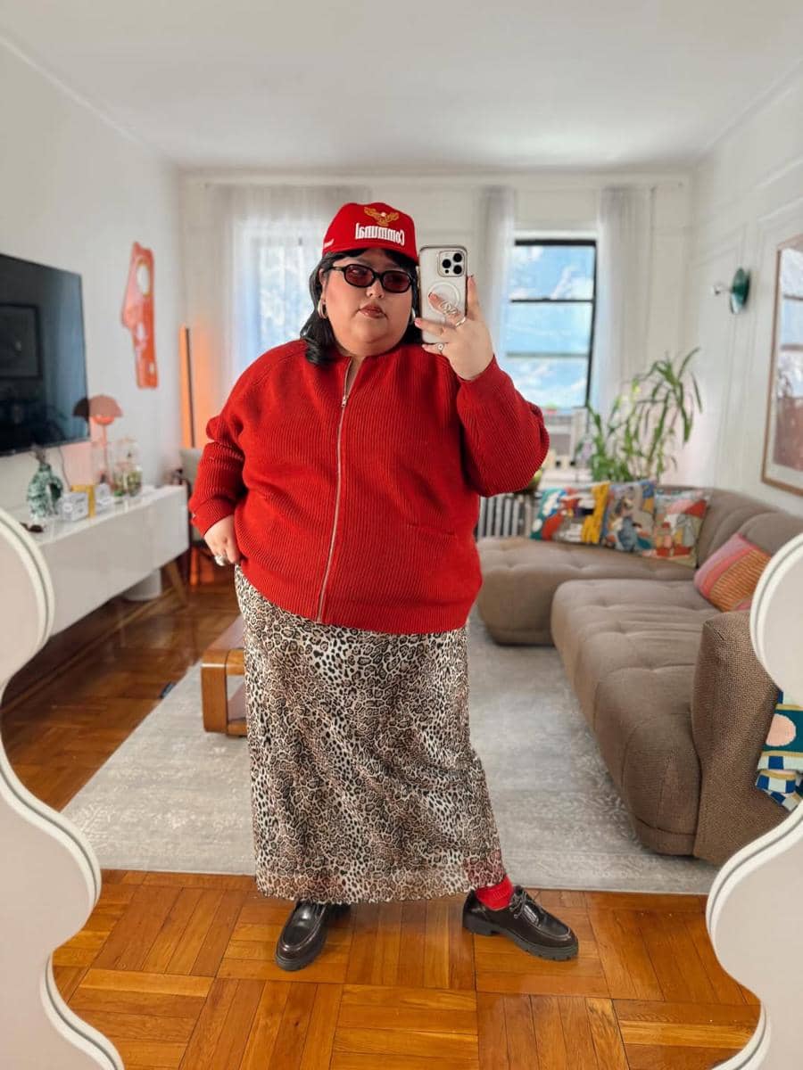 Seorang perempuan berpose di depan cermin mengenakan jaket merah, rok motif leopard, topi merah, dan kacamata hitam di ruang tamu modern.