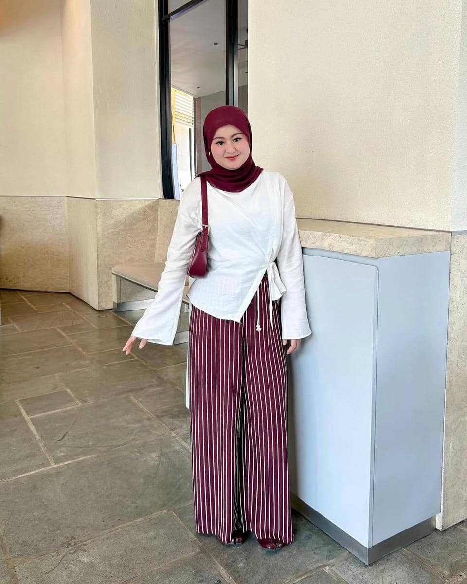 Seorang wanita berhijab merah marun mengenakan blus putih dan celana panjang bergaris vertikal merah marun berdiri di area dalam gedung.