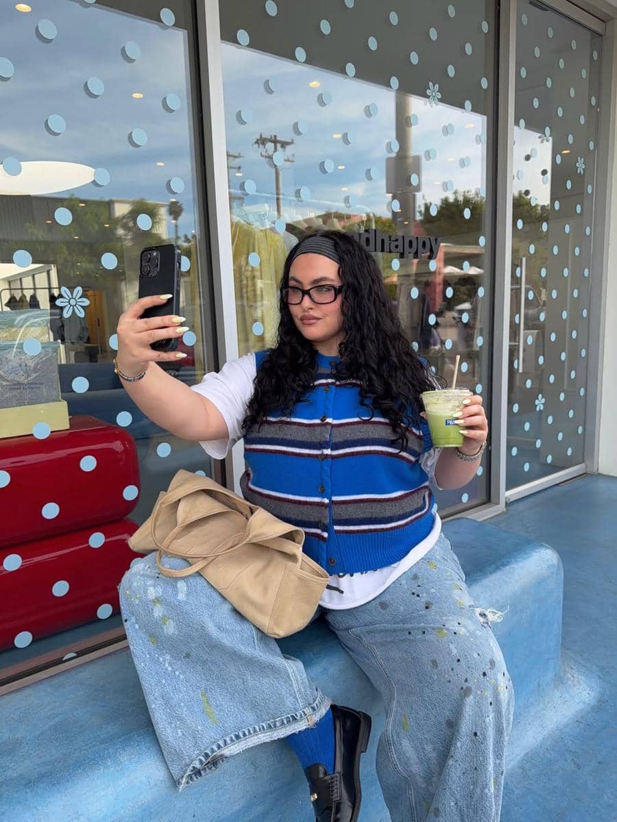 Seorang perempuan berambut panjang mengenakan rompi biru bergaris dan celana jeans longgar sedang berswafoto sambil memegang minuman hijau di depan toko dengan jendela bermotif titik biru.