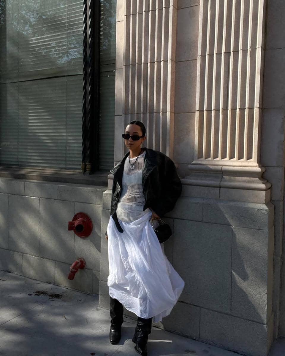 Seorang wanita bergaya street style mengenakan jaket kulit hitam, rok putih panjang, dan kacamata hitam berdiri di depan gedung batu klasik.