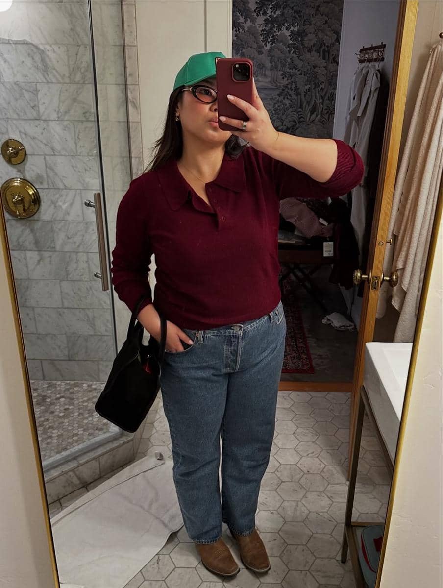 Seorang wanita berpose di depan cermin mengenakan sweter merah marun, celana jeans biru, topi hijau, dan sepatu cokelat di kamar mandi bergaya marmer.
