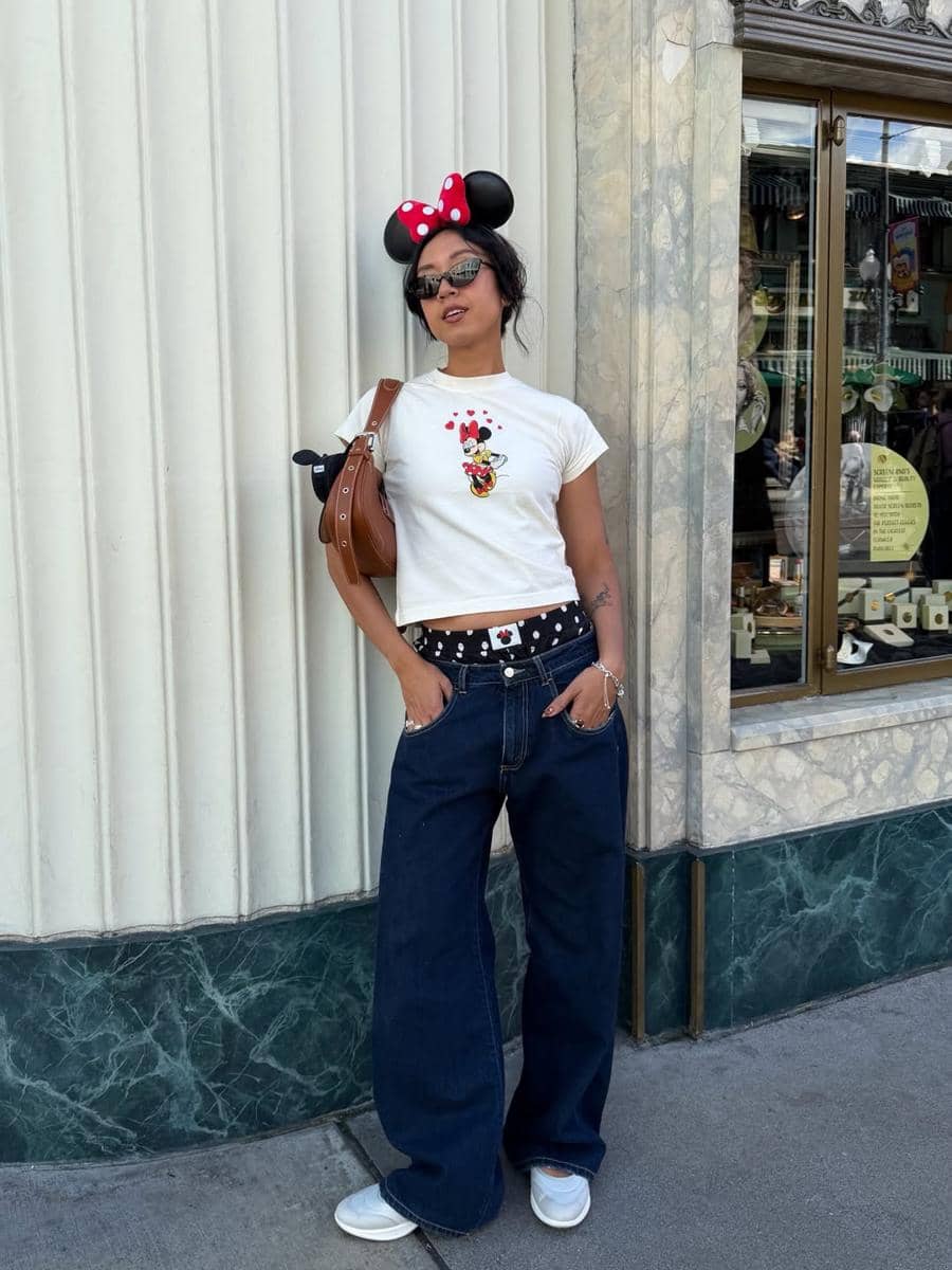 Seorang wanita mengenakan kaus putih bergambar karakter kartun, celana jeans longgar, dan bando telinga Minnie Mouse berdiri di trotoar.