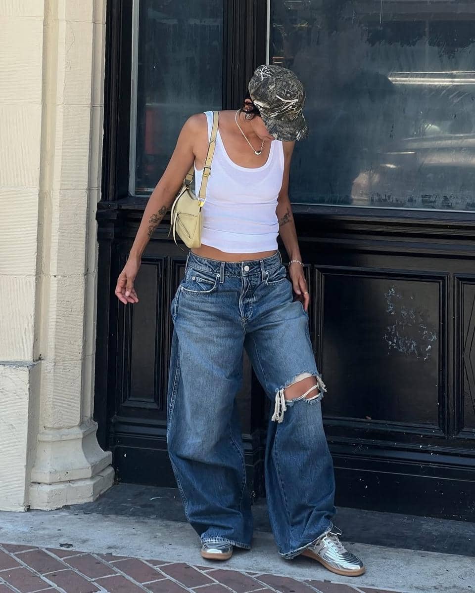 Seseorang mengenakan tank top putih, celana jeans longgar robek di lutut, topi kamuflase, dan tas kecil krem di bahu.