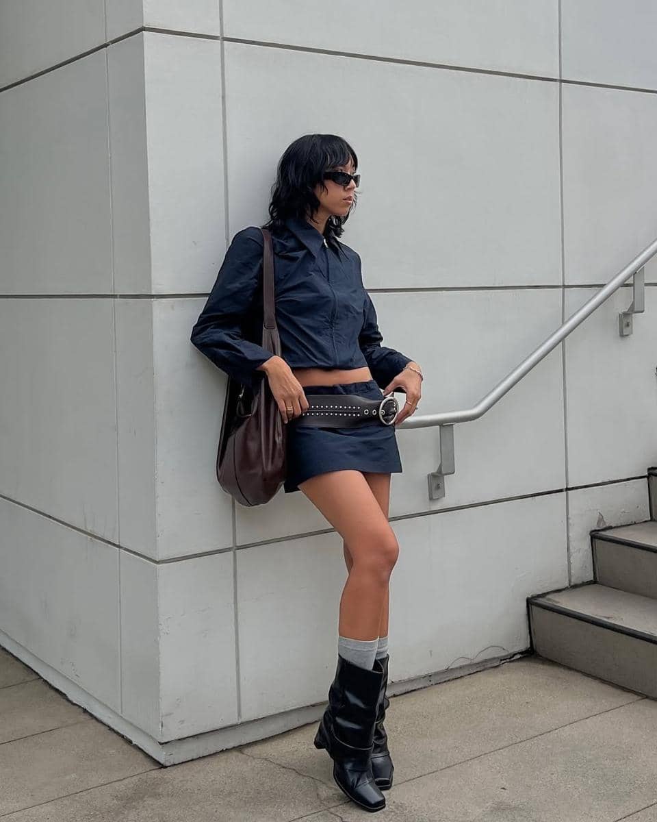 Seorang wanita bergaya dengan kemeja hitam, rok mini, dan sepatu bot hitam berdiri di dekat dinding beton modern sambil mengenakan kacamata hitam.