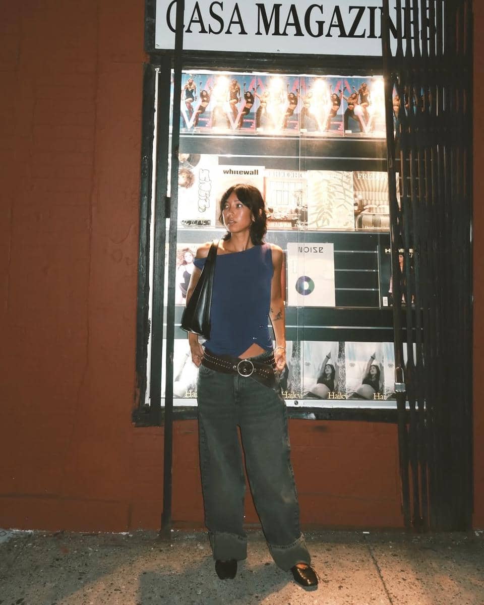 Seorang wanita berdiri di depan etalase bertuliskan CASA MAGAZINE, mengenakan atasan biru, celana jeans longgar, dan tas hitam di bahunya.