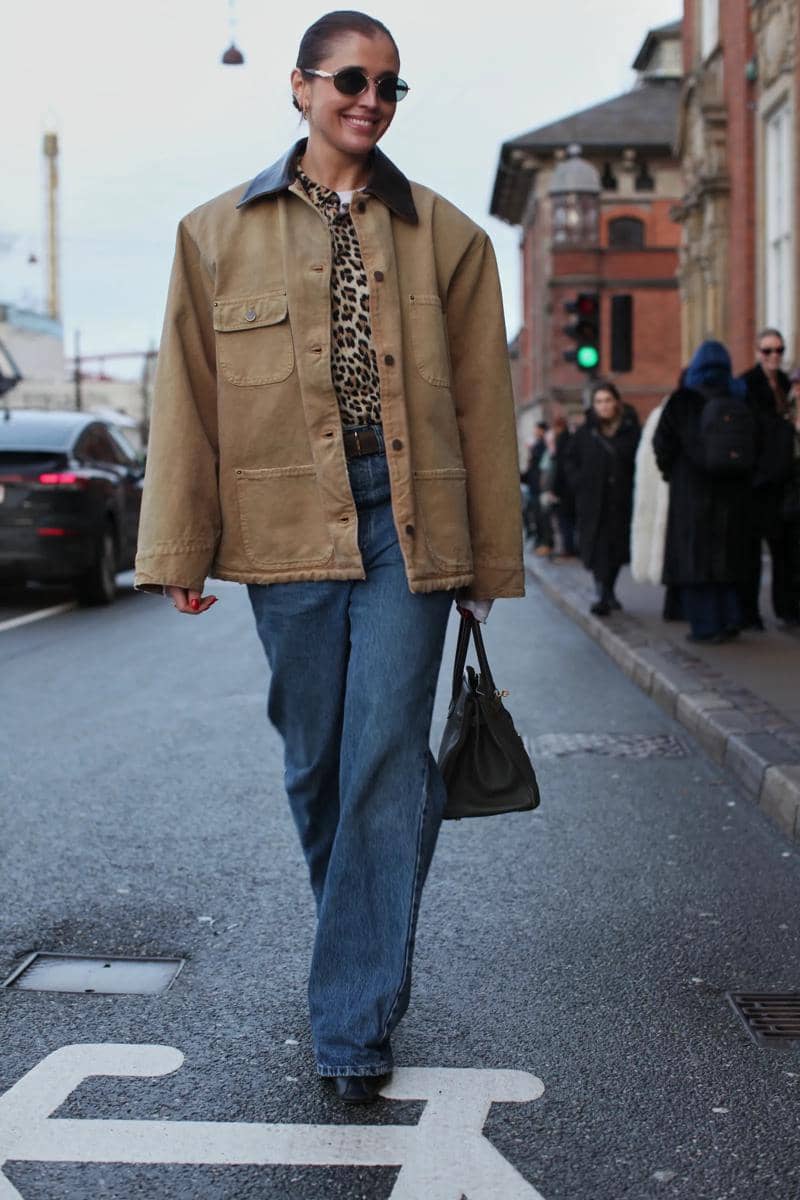 Seorang wanita berjalan di jalan kota mengenakan jaket cokelat oversized, kemeja motif leopard, celana jeans longgar, dan kacamata hitam.