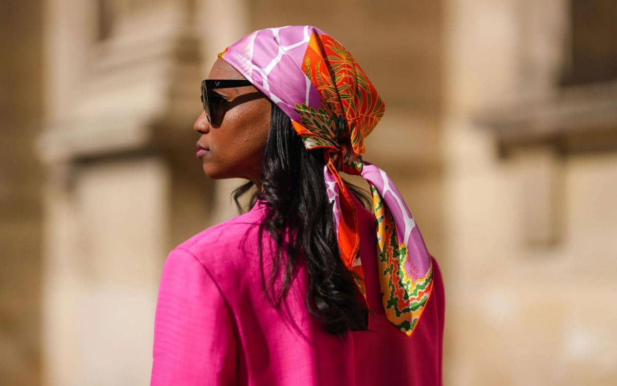 Seorang wanita mengenakan kacamata hitam, blus berwarna fuchsia, dan syal sutra bermotif warna oranye, merah muda, serta hijau di kepala.