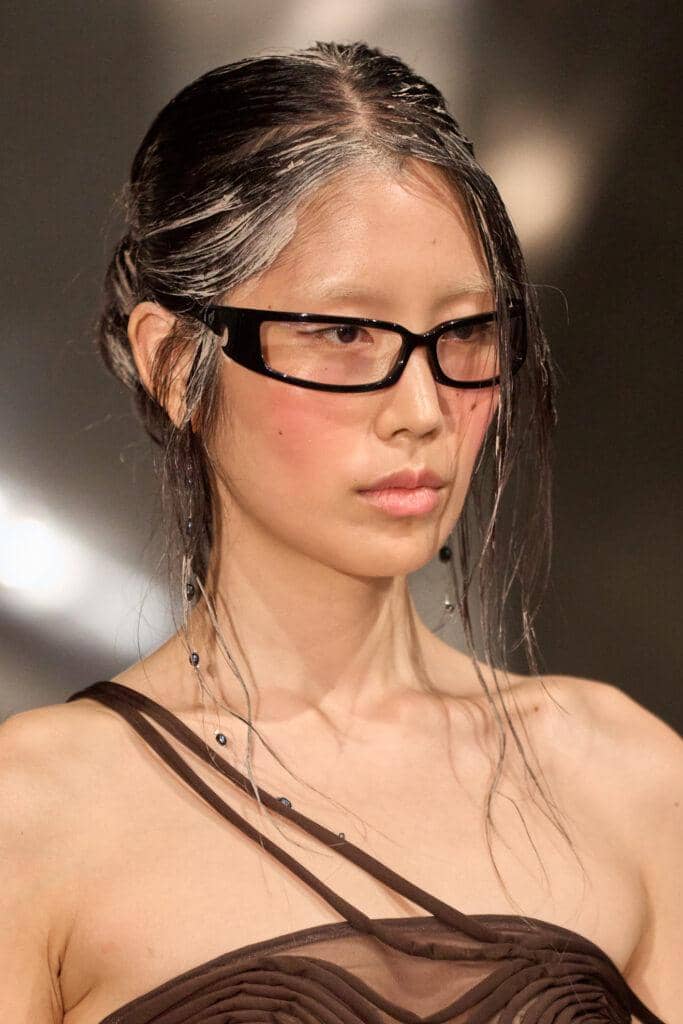 Model mengenakan kacamata hitam bergaya futuristik dengan rambut basah dan gaun cokelat berpotongan unik di atas panggung peragaan busana.
