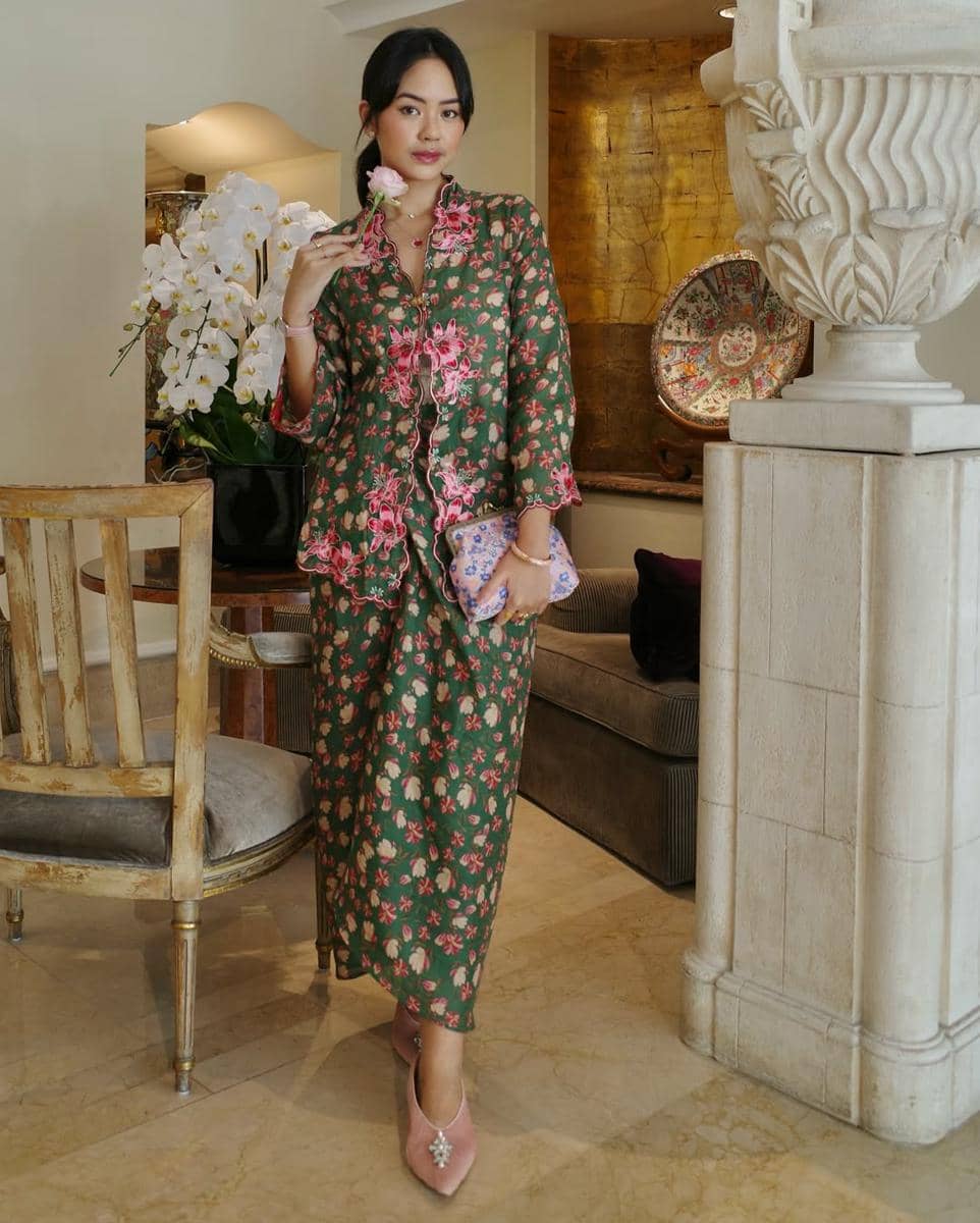 Seorang wanita mengenakan kebaya hijau bermotif bunga merah muda sambil memegang bunga dan tas kecil di ruangan bergaya klasik.