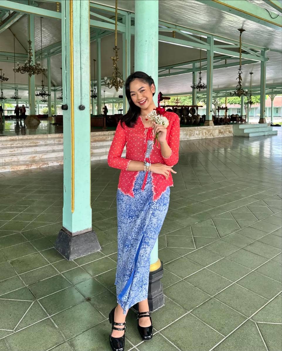 Seorang wanita mengenakan kebaya merah dan kain batik biru berdiri tersenyum sambil memegang bunga di bangunan tradisional Jawa.