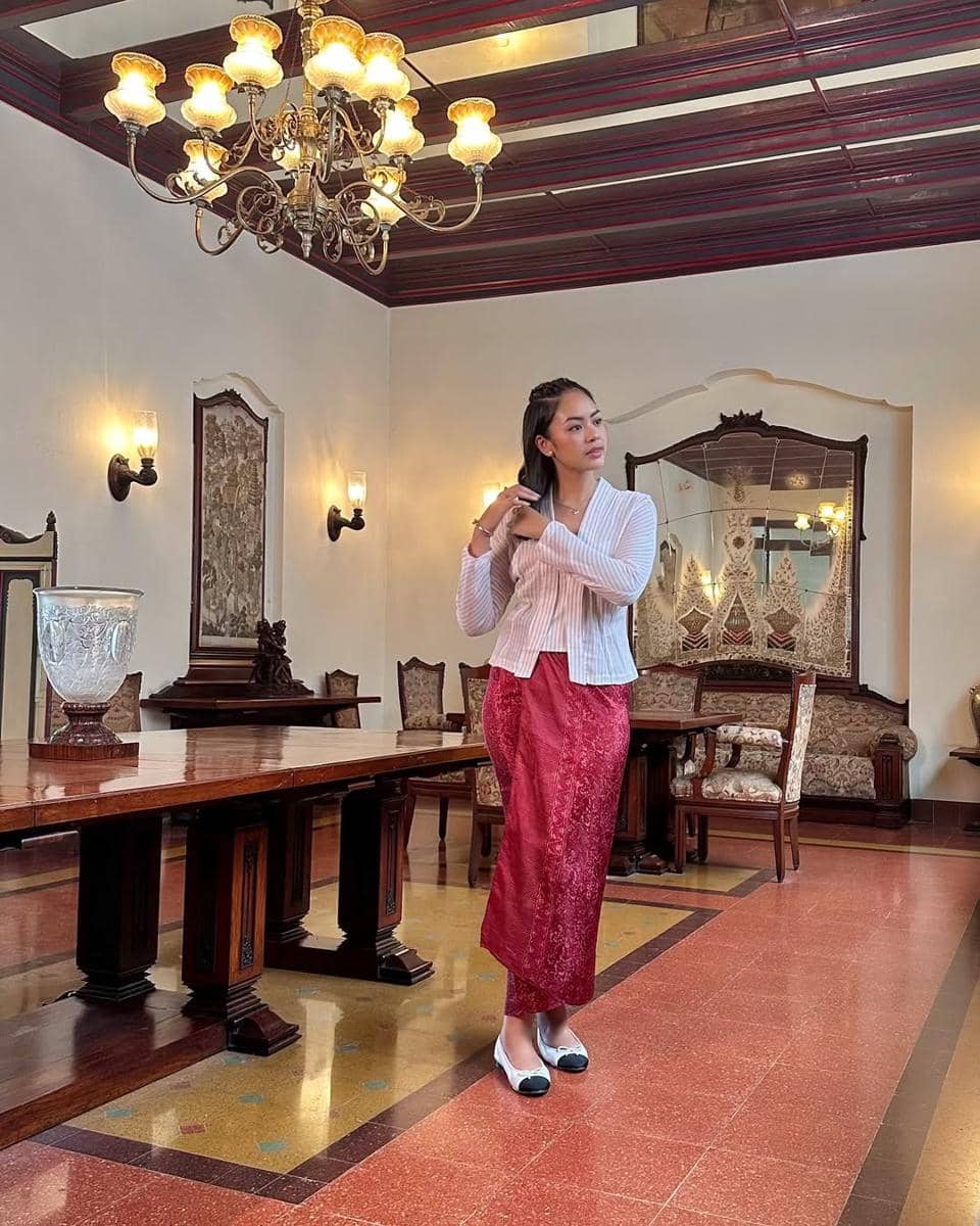 Seorang wanita mengenakan kebaya putih dan kain batik merah berdiri di ruang bergaya klasik dengan lampu gantung dan furnitur kayu antik.