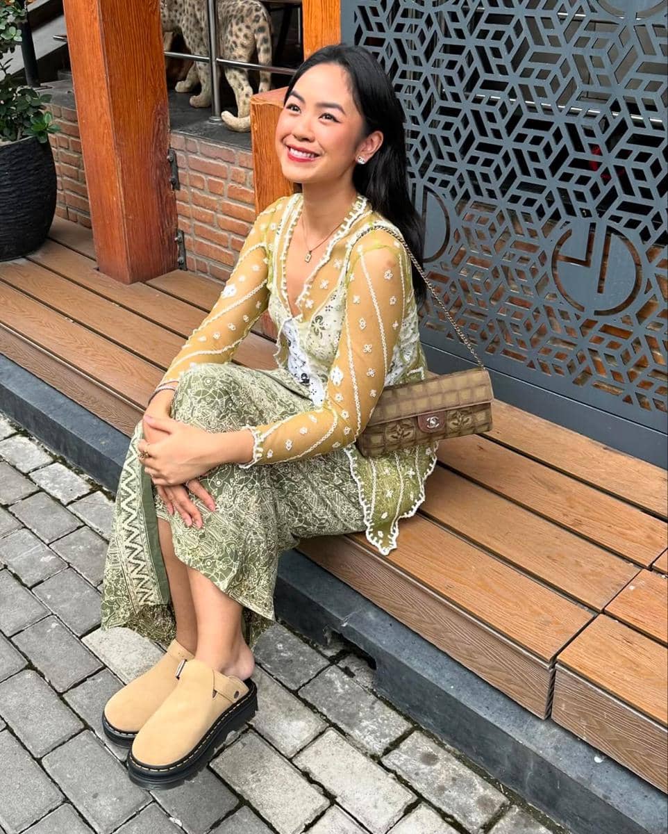 Seorang wanita tersenyum duduk di bangku kayu mengenakan kebaya modern berwarna hijau dan kuning dengan tas cokelat di bahunya.