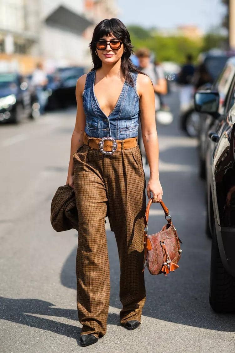 Seorang wanita berjalan di jalan mengenakan rompi denim tanpa lengan, celana panjang motif kotak cokelat, dan membawa tas kulit cokelat.