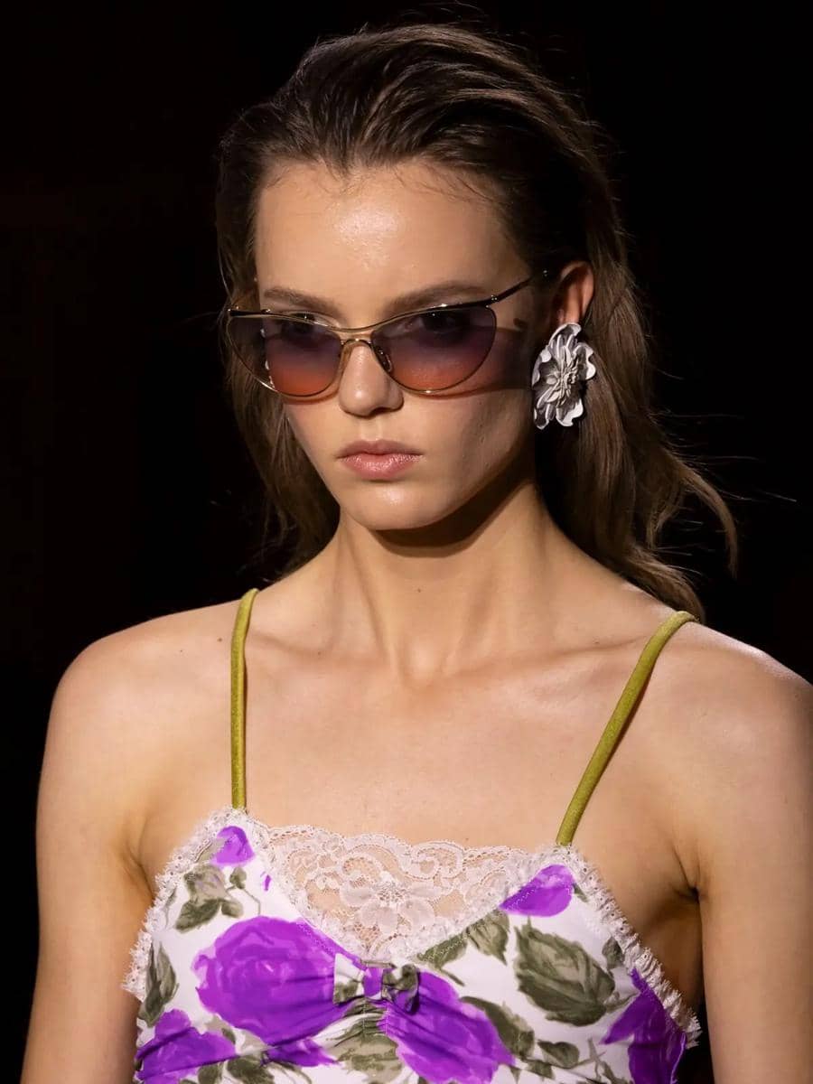 Model mengenakan gaun bermotif bunga ungu dengan renda putih, kacamata hitam gradasi, dan anting besar berbentuk bunga di atas panggung peragaan busana.