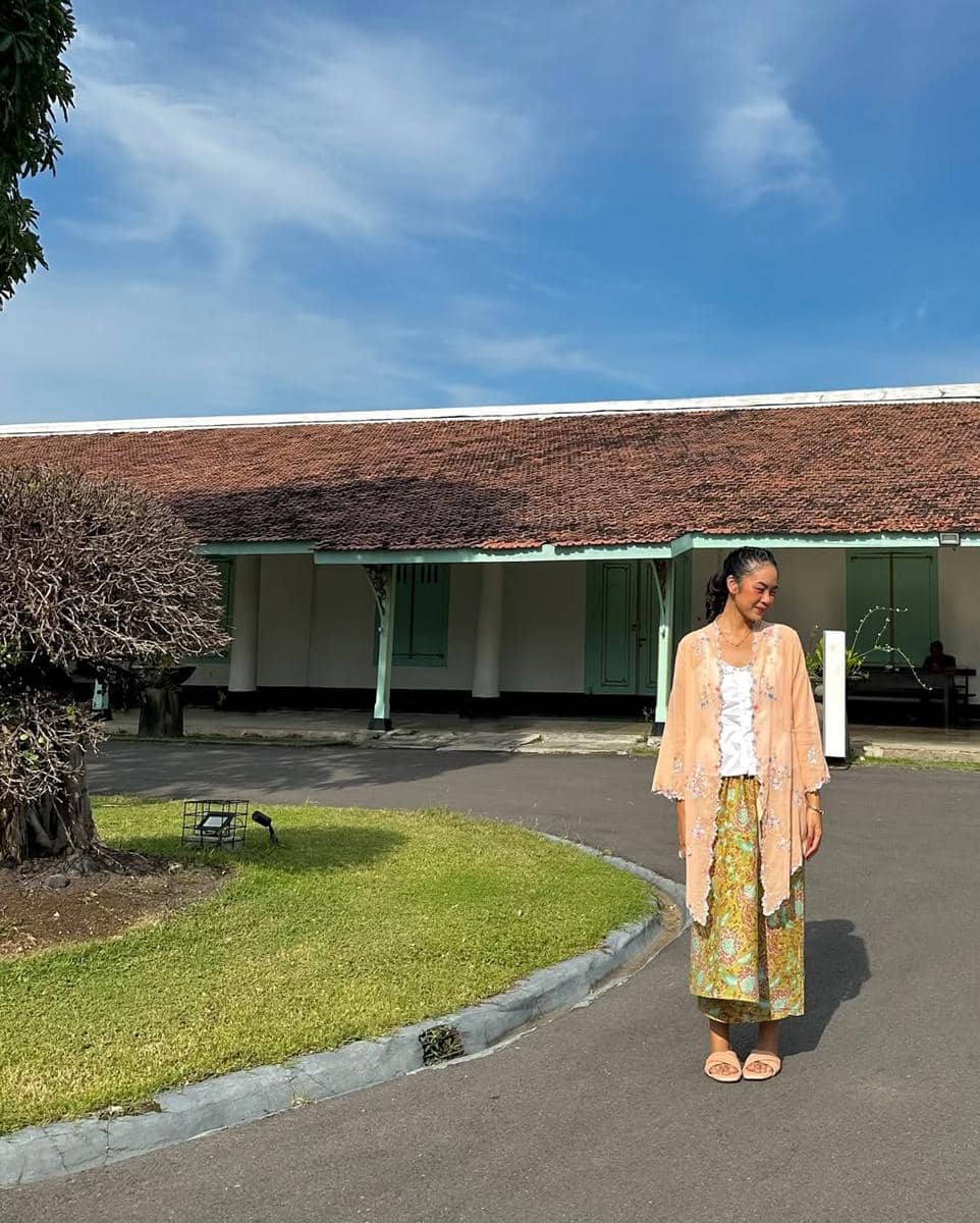 Seorang wanita mengenakan kebaya berwarna peach dan kain batik berdiri di halaman bangunan bergaya kolonial dengan langit biru cerah.