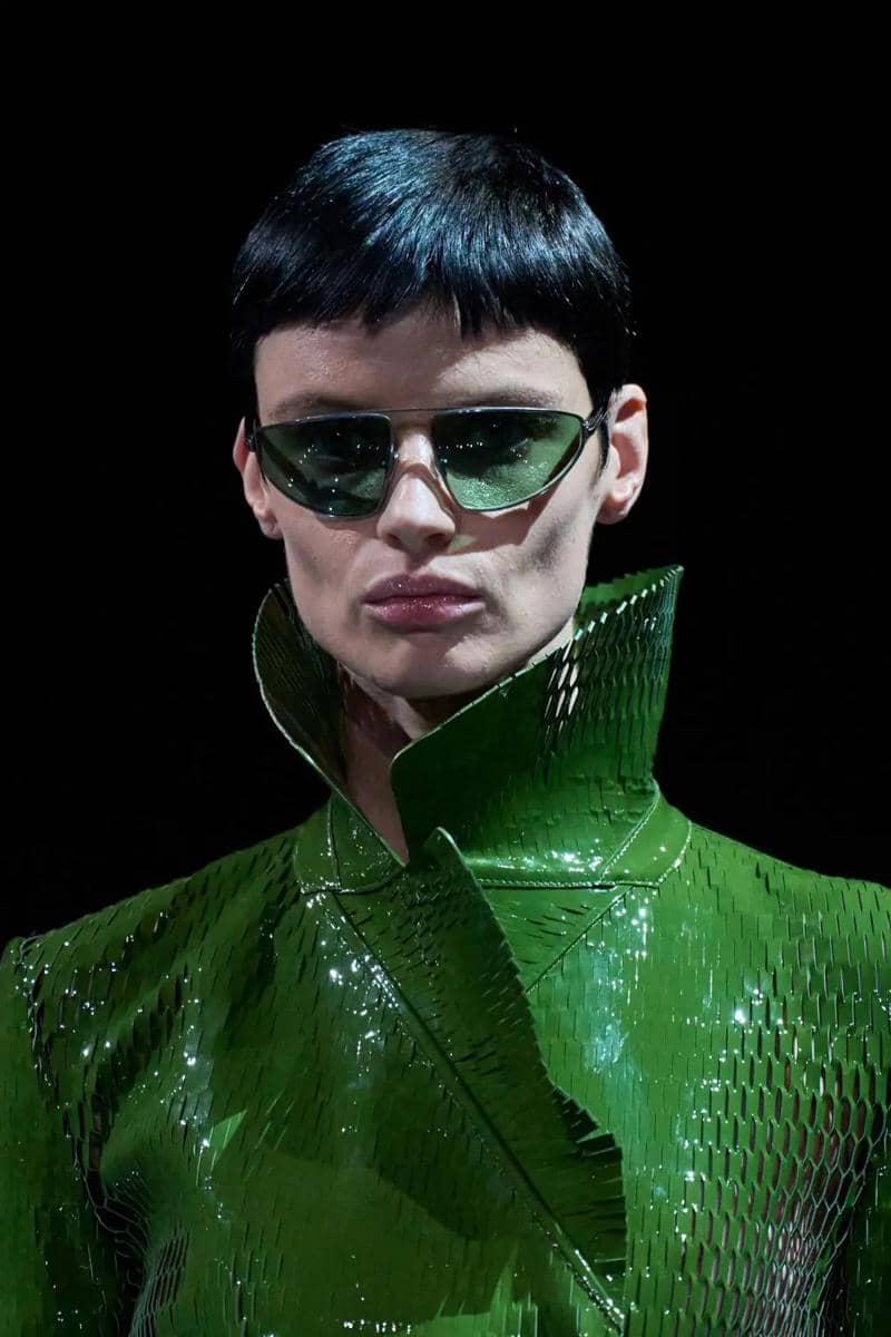 Model berambut pendek mengenakan jaket hijau metalik bertekstur dan kacamata hitam hijau dengan latar belakang gelap.
