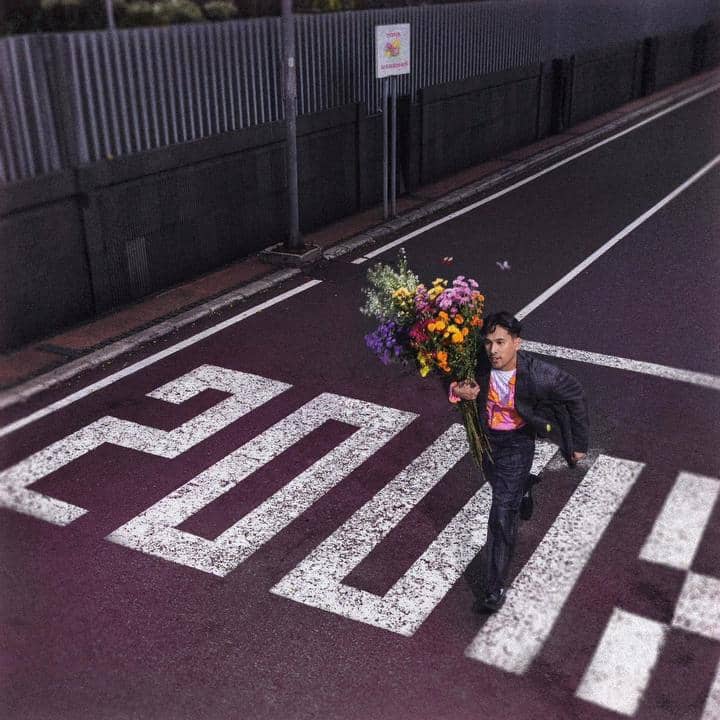 Seorang pria mengenakan jas hitam berjalan di zebra cross sambil membawa buket besar bunga berwarna-warni di jalan kota yang sepi.