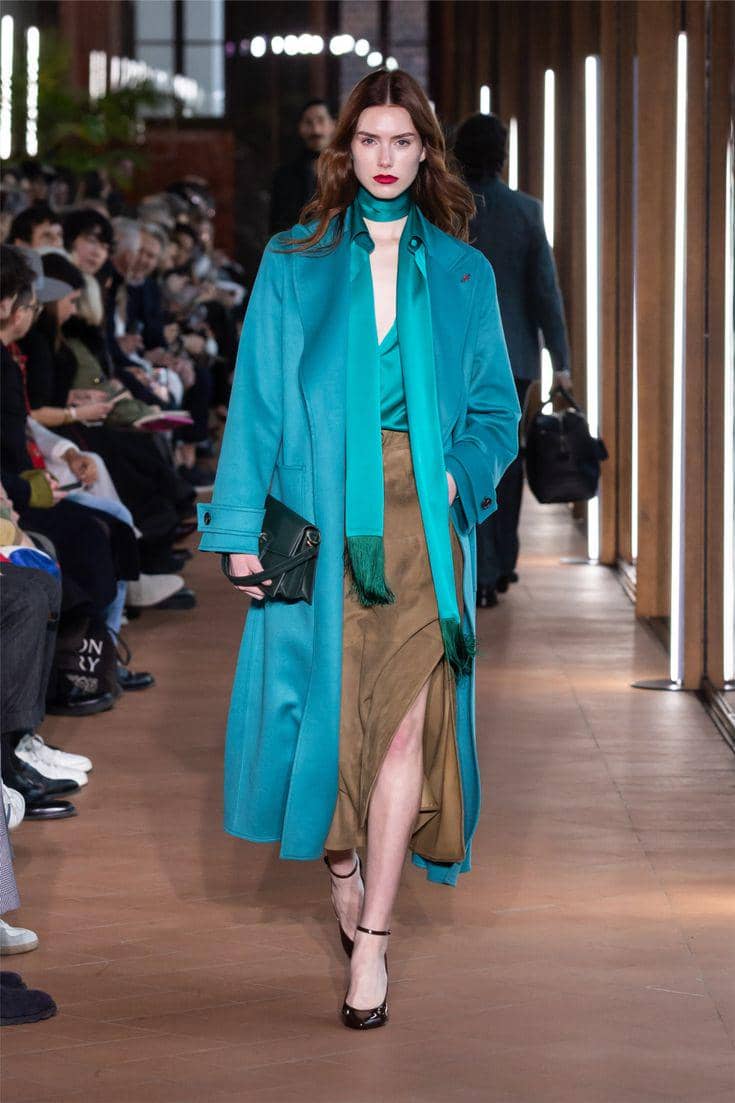 Model berjalan di atas runway mengenakan mantel biru toska panjang, rok cokelat belahan tinggi, dan syal hijau di acara fashion show.