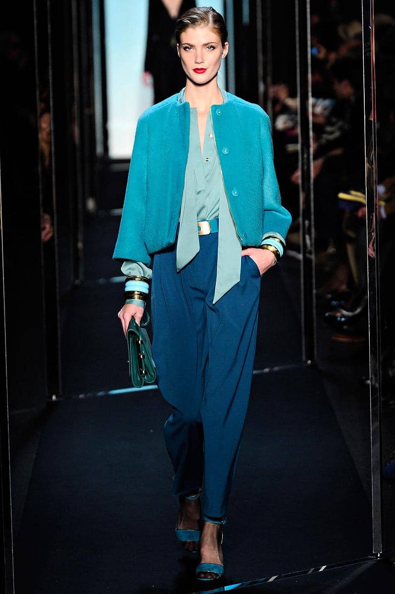 Model berjalan di atas runway mengenakan busana bernuansa biru toska dengan celana longgar, blus, dan jaket pendek serasi.
