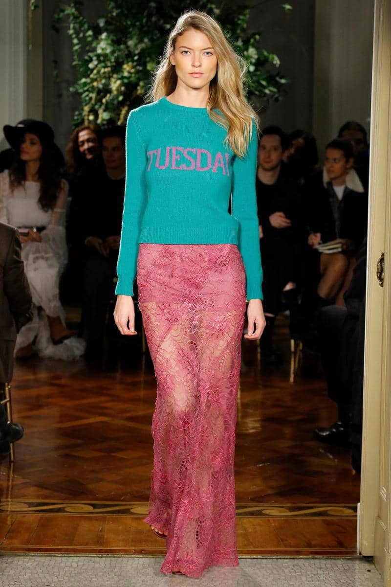 Model berjalan di runway mengenakan sweater hijau bertuliskan 'Tuesday' dan rok panjang renda berwarna merah muda transparan.