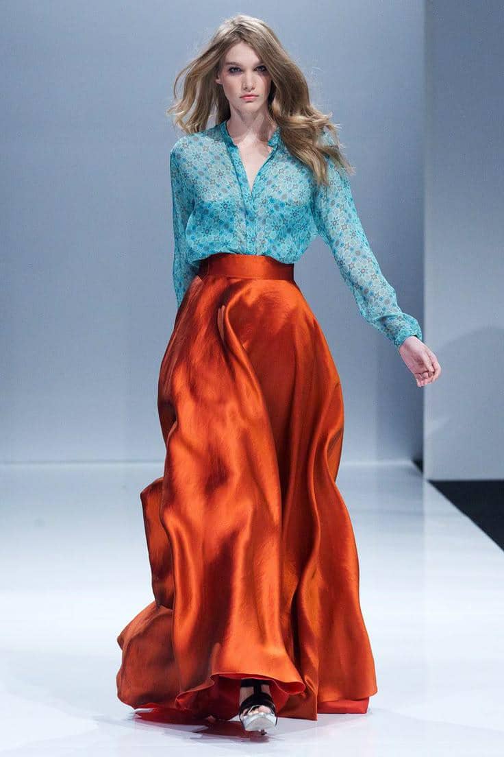 Model berjalan di runway mengenakan blus biru bermotif renda dan rok panjang satin berwarna oranye mengilap.