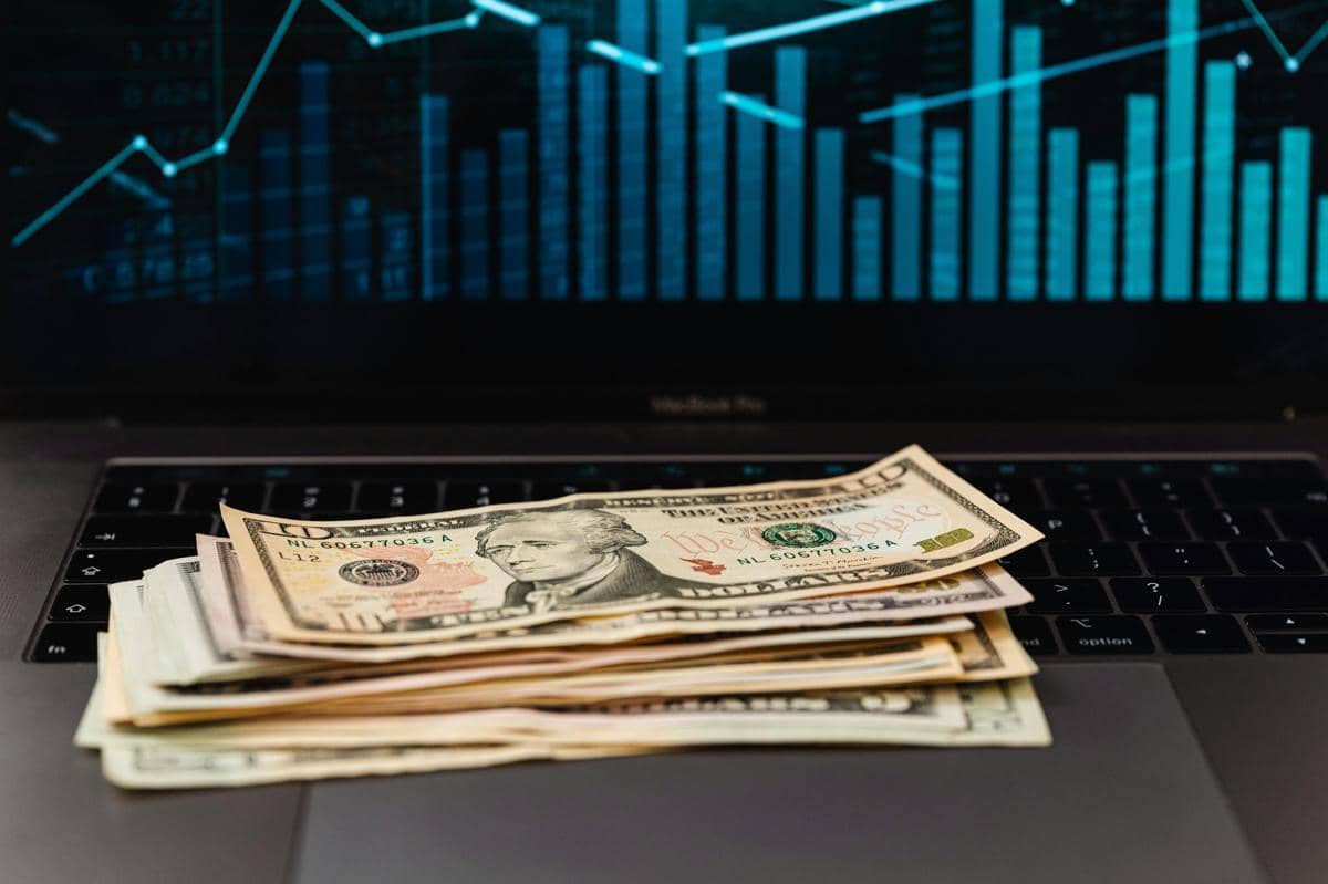 Tumpukan uang dolar Amerika diletakkan di atas keyboard laptop dengan grafik saham biru di layar