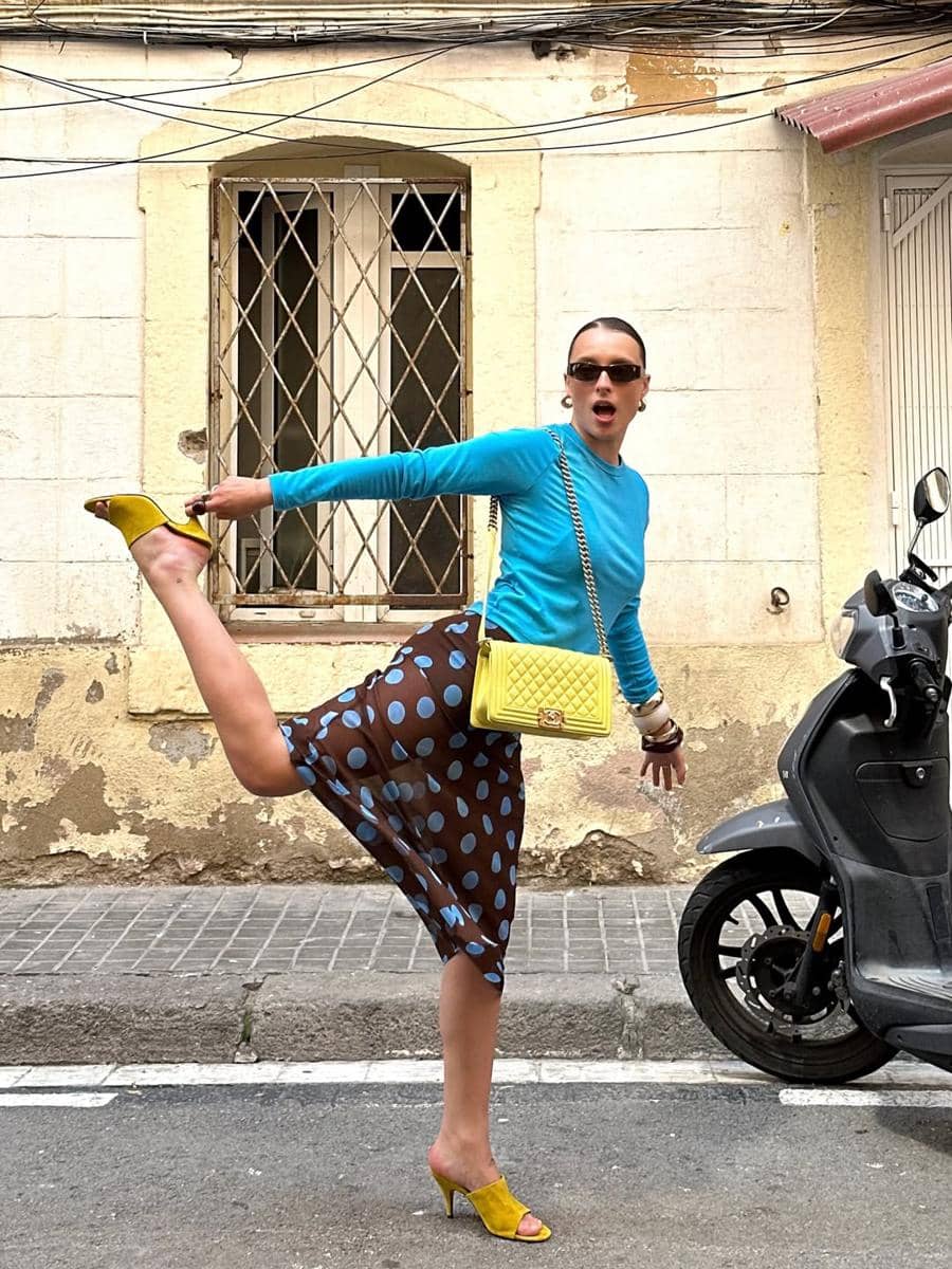 Seorang wanita bergaya di jalan dengan mengenakan atasan biru, rok transparan bermotif polkadot, tas kuning, dan sepatu hak tinggi kuning.