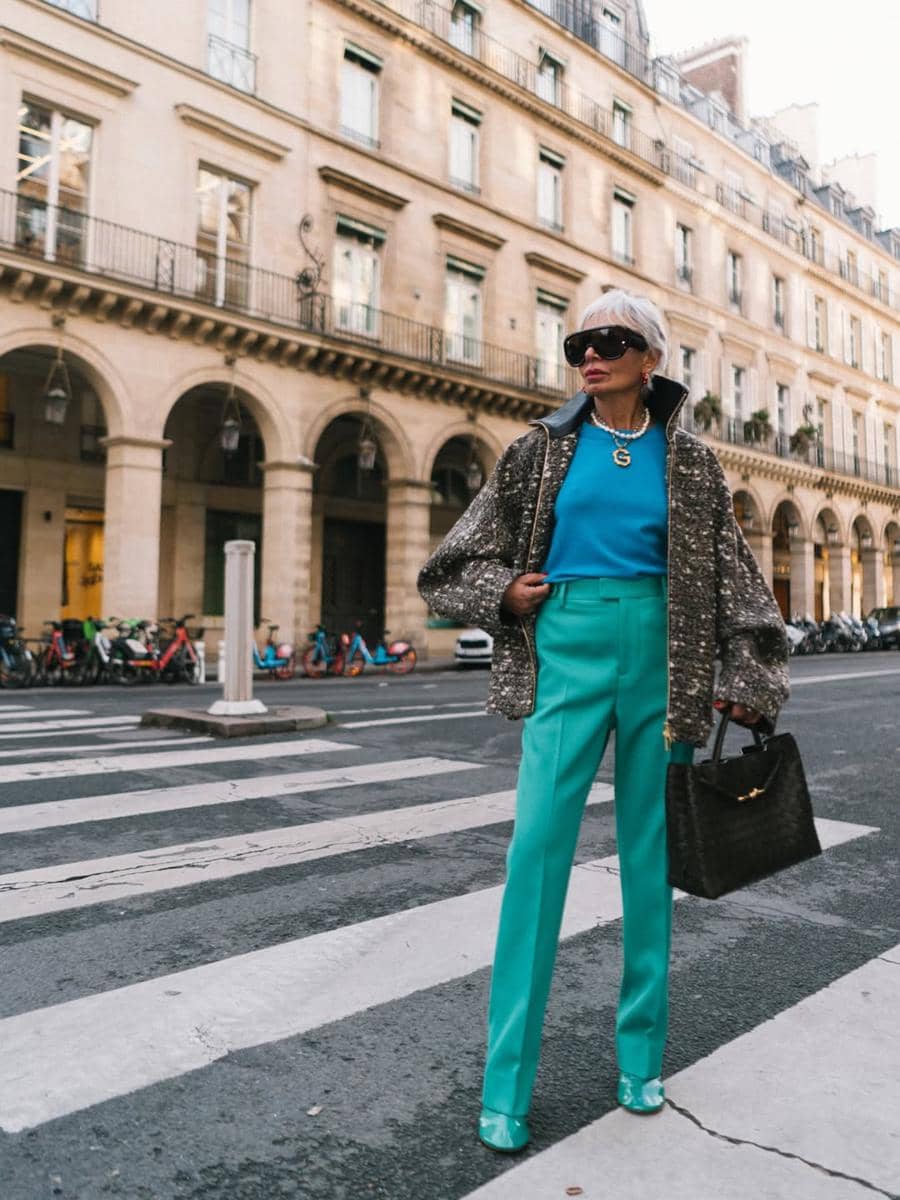 Seorang wanita bergaya elegan mengenakan celana panjang hijau toska, atasan biru, dan jaket bermotif di jalan kota dengan latar bangunan klasik.