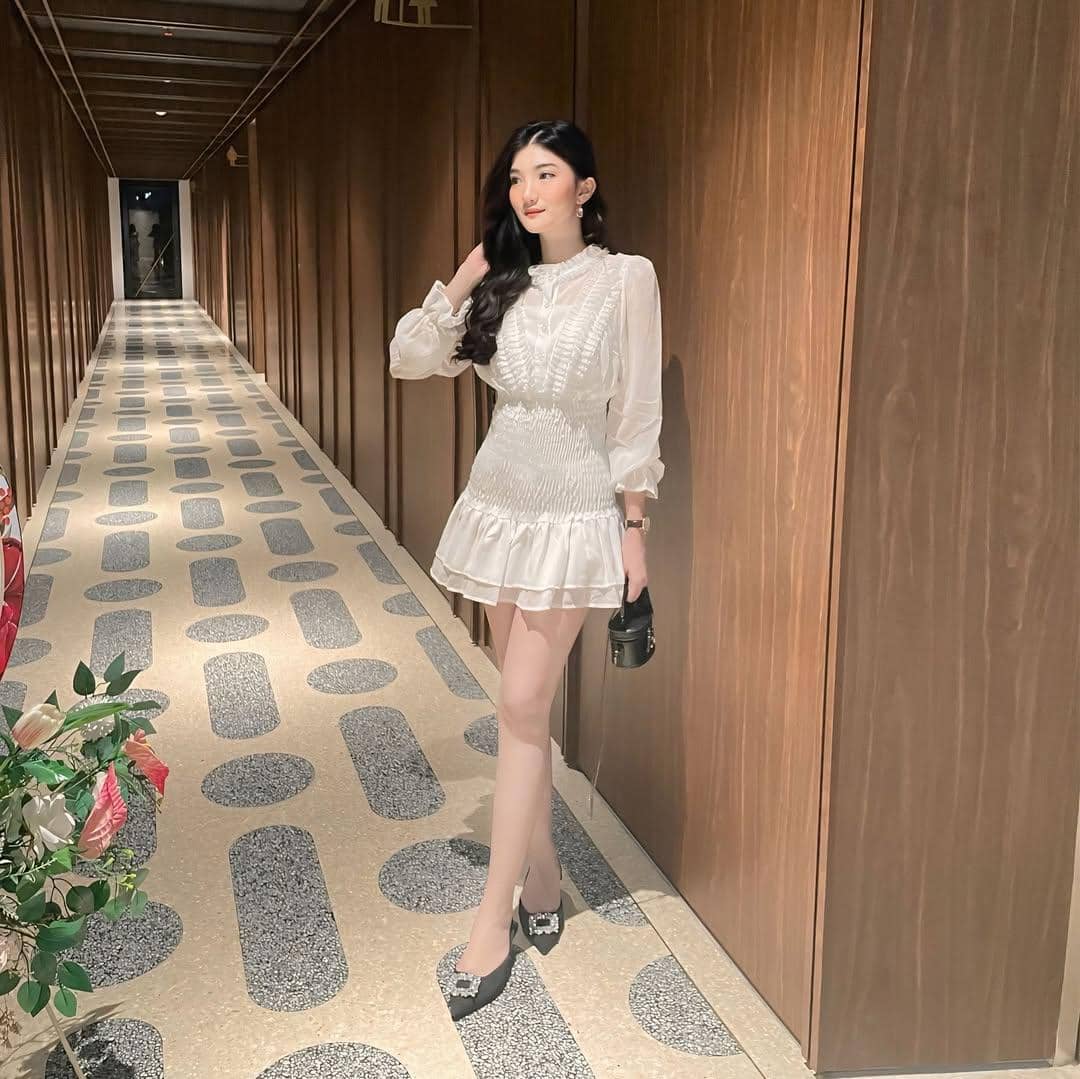 Seorang wanita mengenakan gaun putih lengan panjang berdiri di lorong dengan dinding kayu dan lantai bermotif geometris.