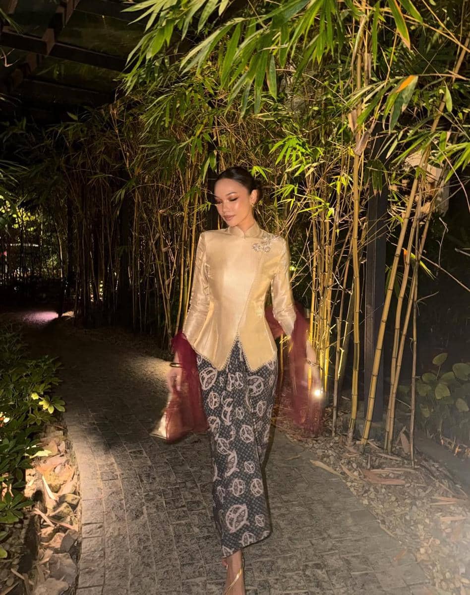 Seorang wanita mengenakan kebaya modern berwarna emas dengan lengan renda merah dan kain batik berjalan di jalan setapak di taman bambu pada malam hari.