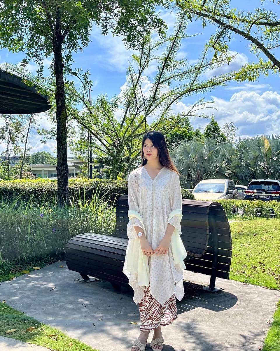 Seorang wanita berdiri di taman dengan latar pepohonan hijau dan langit biru cerah, mengenakan pakaian berwarna putih dan cokelat bermotif.
