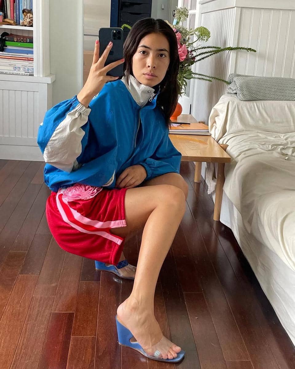 Seorang wanita berpose di dalam ruangan sambil berfoto di cermin, mengenakan jaket biru, celana pendek merah, dan sandal hak biru transparan.