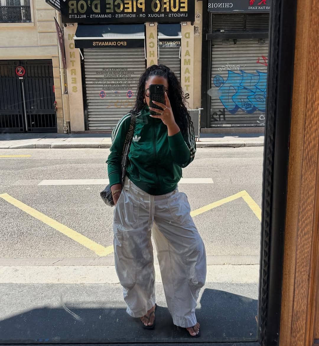 Seorang wanita berpose di depan cermin jalanan mengenakan jaket hijau Adidas dan celana putih longgar sambil memegang ponsel.