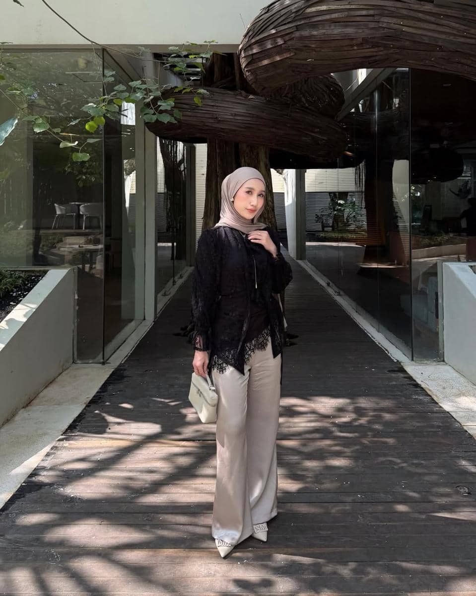 Seorang wanita berhijab mengenakan atasan hitam dan celana panjang krem berpose di area luar ruangan dengan latar arsitektur modern dan pepohonan.