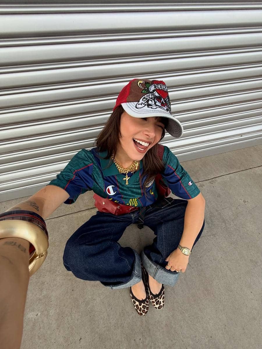 Seorang perempuan tersenyum sambil berpose duduk di depan pintu logam, mengenakan topi merah, kemeja hijau, celana jeans longgar, dan sepatu motif leopard.