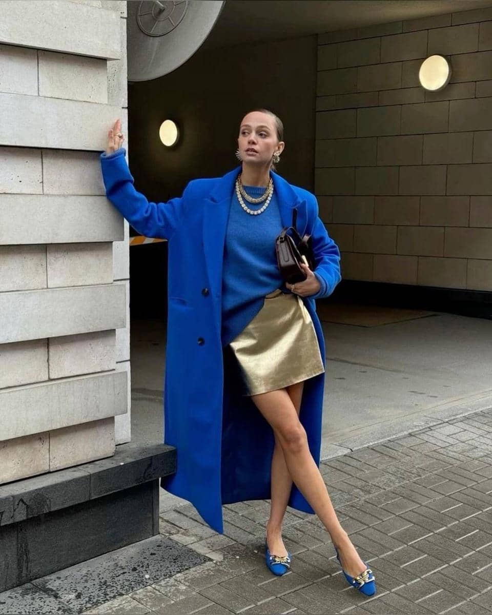 Seorang wanita bergaya mengenakan mantel biru panjang, sweater biru, rok mini emas, dan sepatu hak rendah biru di area perkotaan.