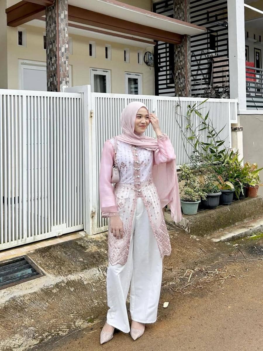 Seorang wanita berhijab mengenakan kebaya modern berwarna pink muda dan celana putih berdiri di depan pagar rumah berwarna putih.