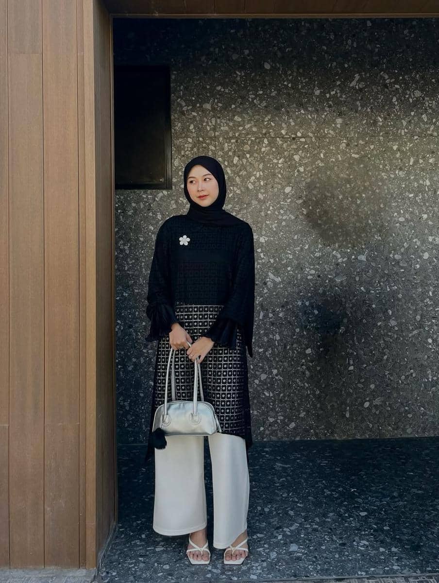 Seorang wanita berhijab hitam mengenakan tunik hitam bermotif dan celana panjang putih, memegang tas tangan berwarna krem di depan dinding batu abu-abu.