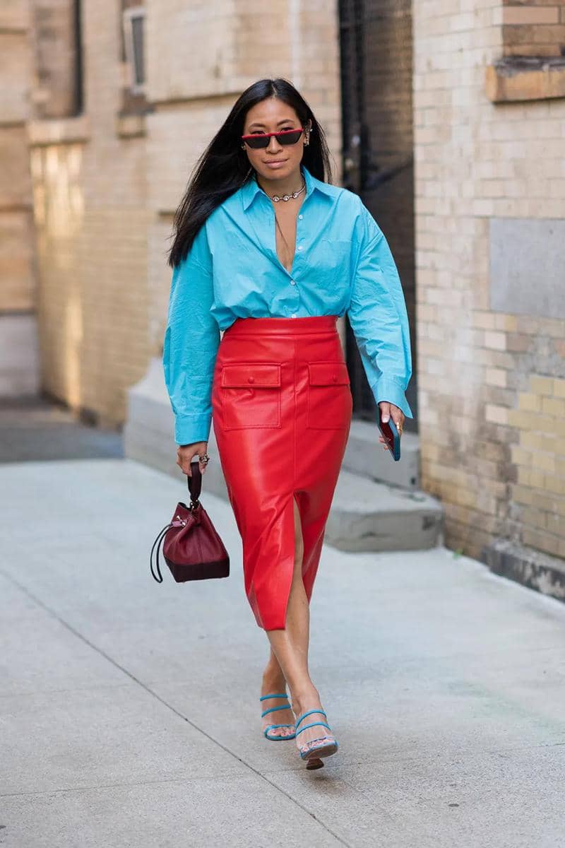 Seorang wanita berjalan di trotoar mengenakan kemeja biru cerah dan rok kulit merah dengan belahan depan, membawa tas kecil merah marun.